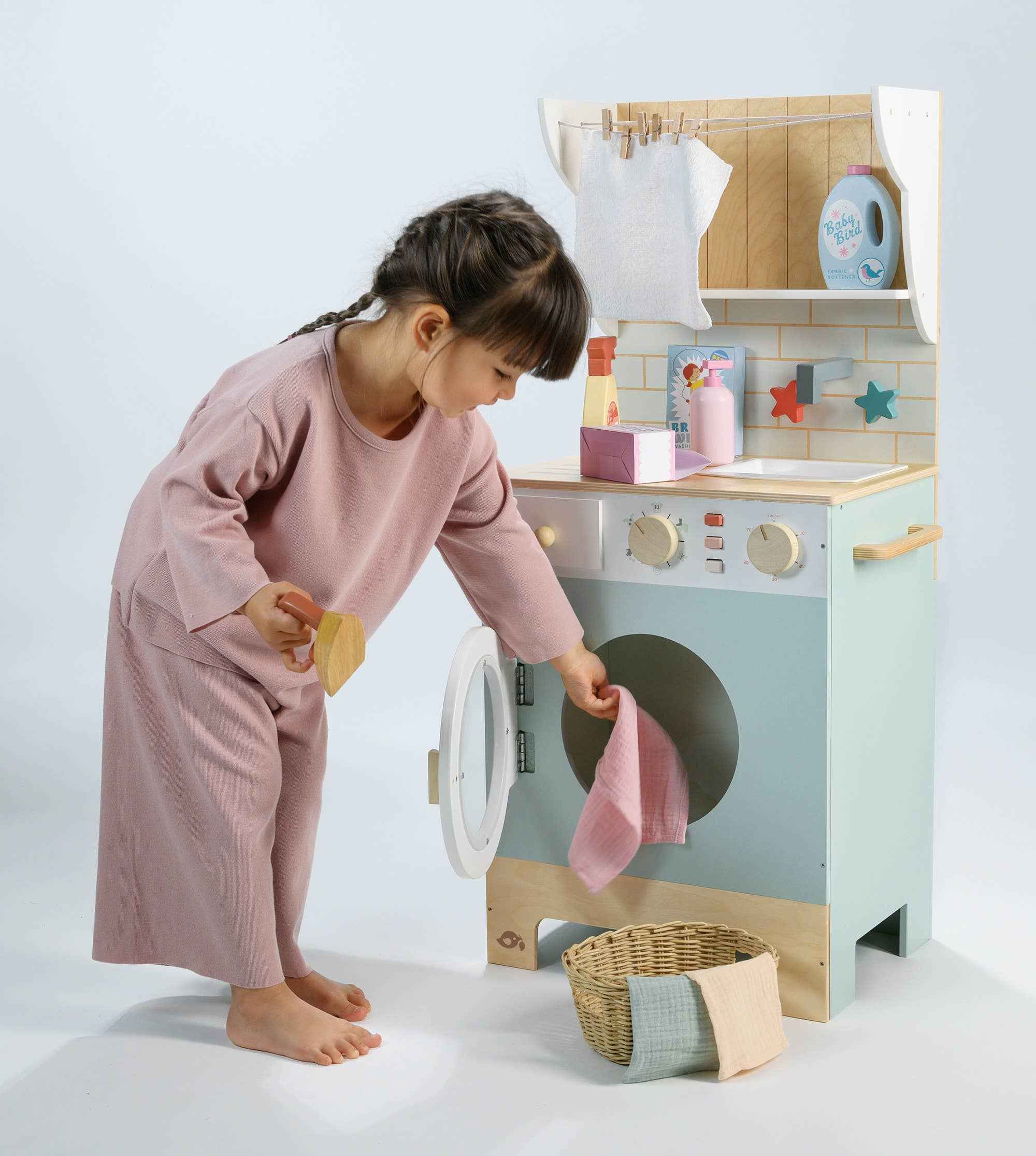 Child playing with Tender Leaf Wooden Laundrette, loading fabric into the toy washing machine in a pretend housekeeping setup.