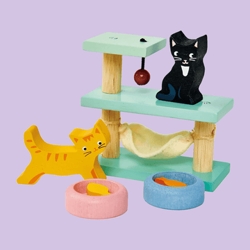 Colorful wooden cat playset featuring two toy cats (one black, one orange), a multi-level cat tree with a hammock and hanging toy, and two food bowls with fish pieces on a purple background.