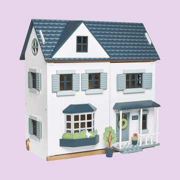 White and blue wooden dollhouse with two stories, bay window, front porch, and decorative plants, set against a light purple background.