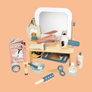 Children's wooden vanity playset with mirror, toy hairdryer, straightener, comb, scissors, lotion bottles, accessories, and a pretend beauty magazine on a peach background.