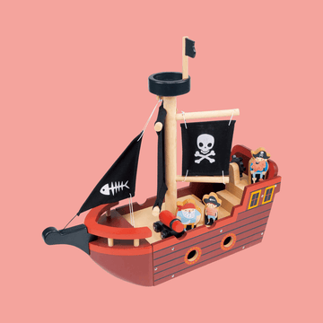 Wooden toy pirate ship with black sails featuring skull and fishbone designs, red and brown hull, and small pirate figurines on deck against a pink background.