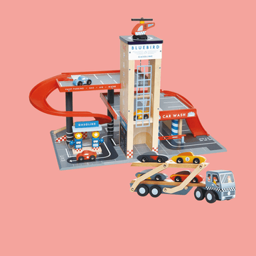 Wooden toy car garage playset featuring a multi-level parking structure, red spiral ramp, gas station, car wash, and a car carrier truck with colorful toy cars on a pink background.