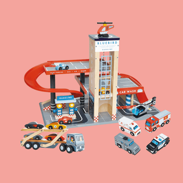 Wooden toy car garage playset with a red spiral ramp, gas station, car wash, elevator tower, and various toy vehicles including cars, trucks, ambulance, and a helicopter on a pink background.